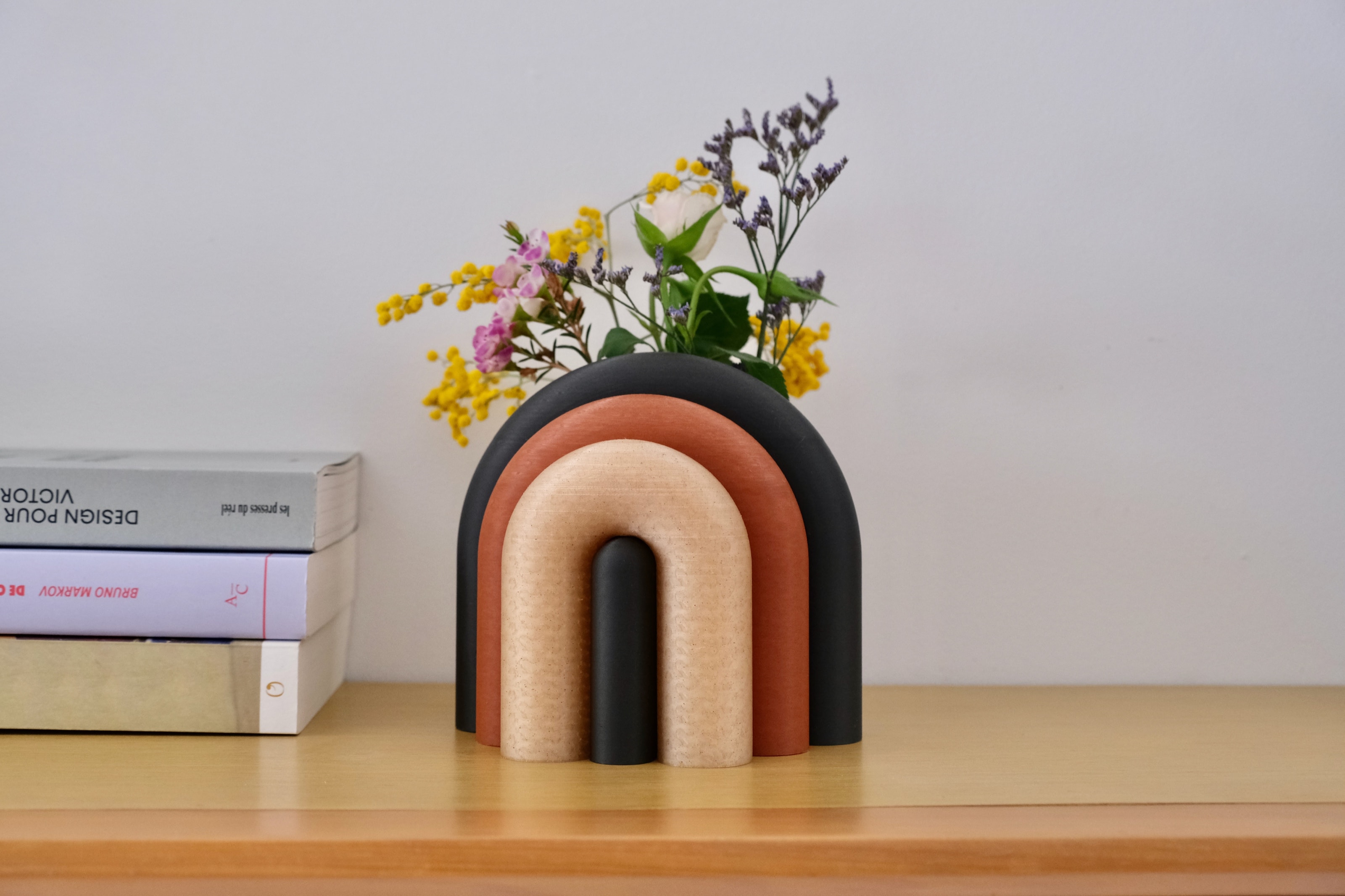 Vase Arc noir, terracotta et bois clair, garni d’un bouquet, posé près d’une pile de livres de design