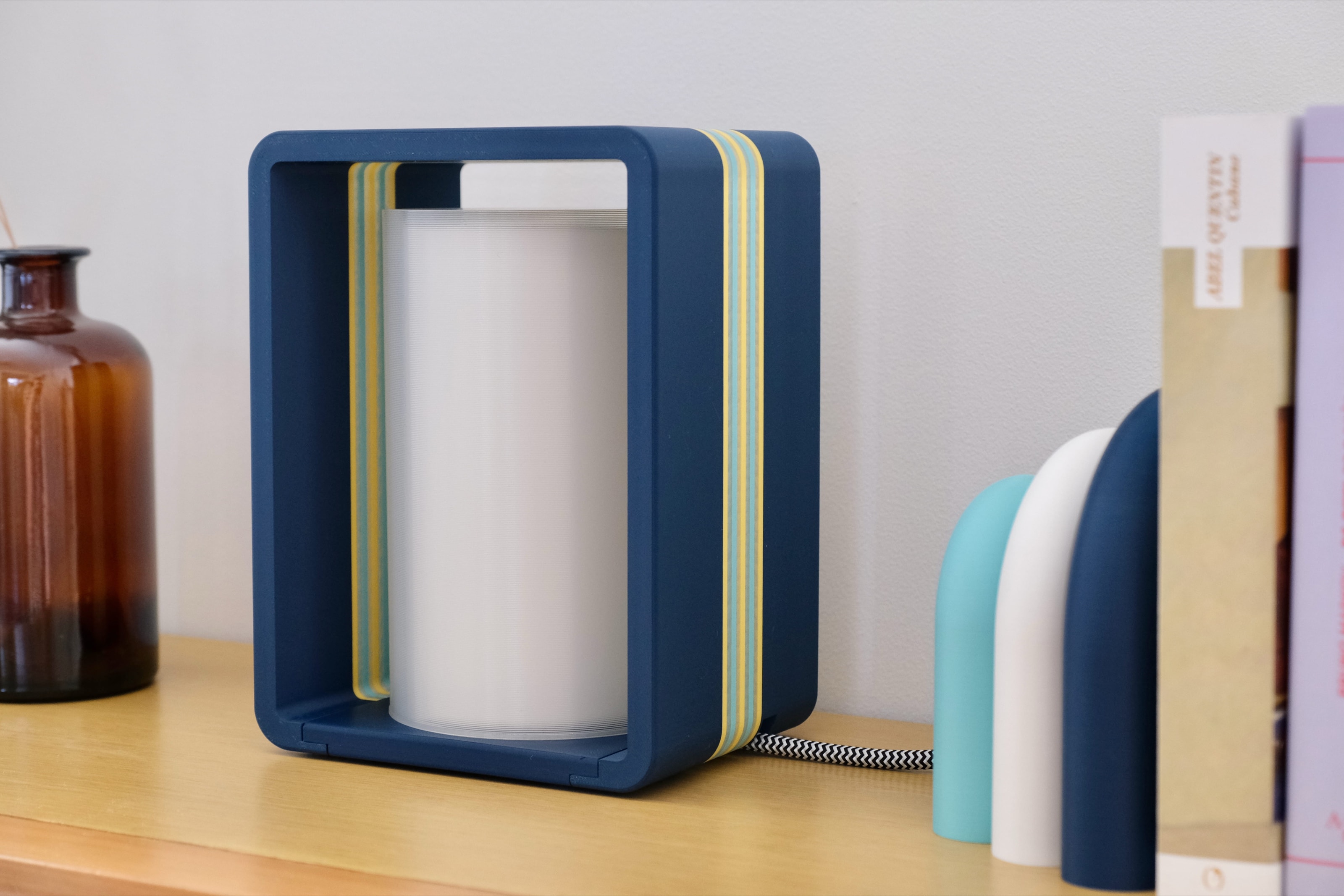 Lampe à poser Interstice bleu marine à liserés jaune et turquoise, diffuseur cylindrique blanc, sur un plan de bois